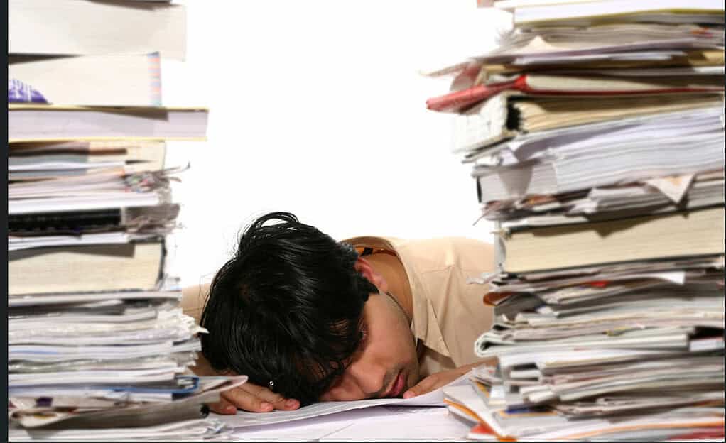head down on desk surrounded by huge piles of papers on both sides of face